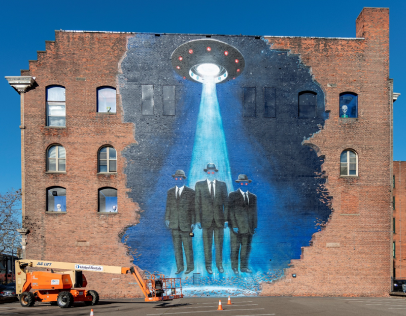 An image of the "Men In Black" mural done by artist Brad Noble. It depicts three faceless men in black suits floating together towards a UFO. The men have glowing red eyes. 