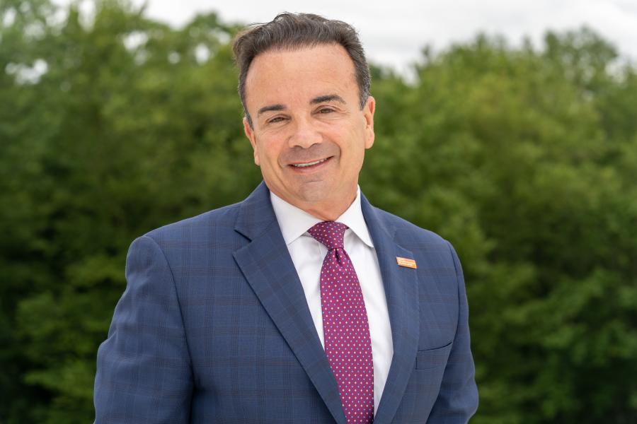 Mayor Ganim posing for a head shot outside with tress in background, while wearing a navy blue blazer and maroon-colored tie