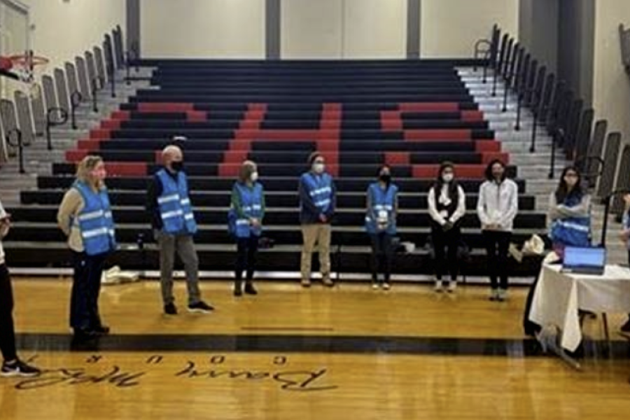 Volunteers in face masks and blue vests stand in a semi-circle in a school gym next to bleachers