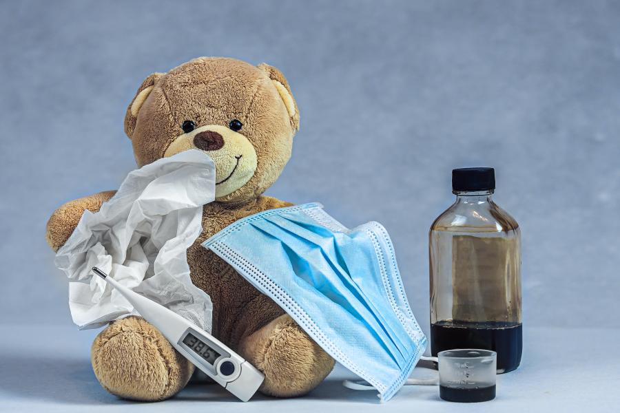 A photo of a brown teddy bear with a surgical mask, tissue, and thermometer on it next to a glass bottle and medicine cup
