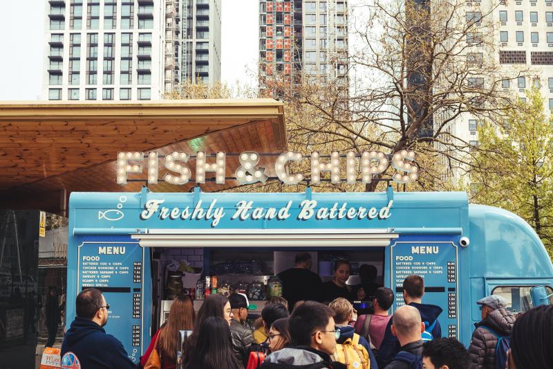 Photo of a blue food truck with lighted Fish and Chips sign on roof and a crowd of people waiting in line