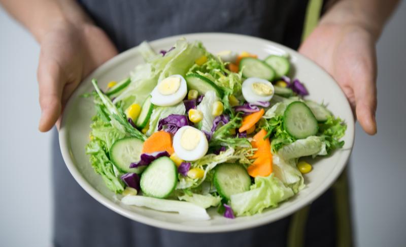 Hands holding out a white bowl containing a fresh salad with lettuce, hardboiled eggs, cucumbers, and carrots