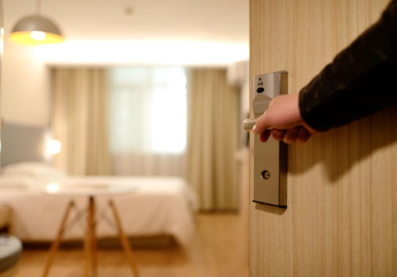 Close up of a person opening a hotel room door with the room in the background including a bed, bright window, and table. 