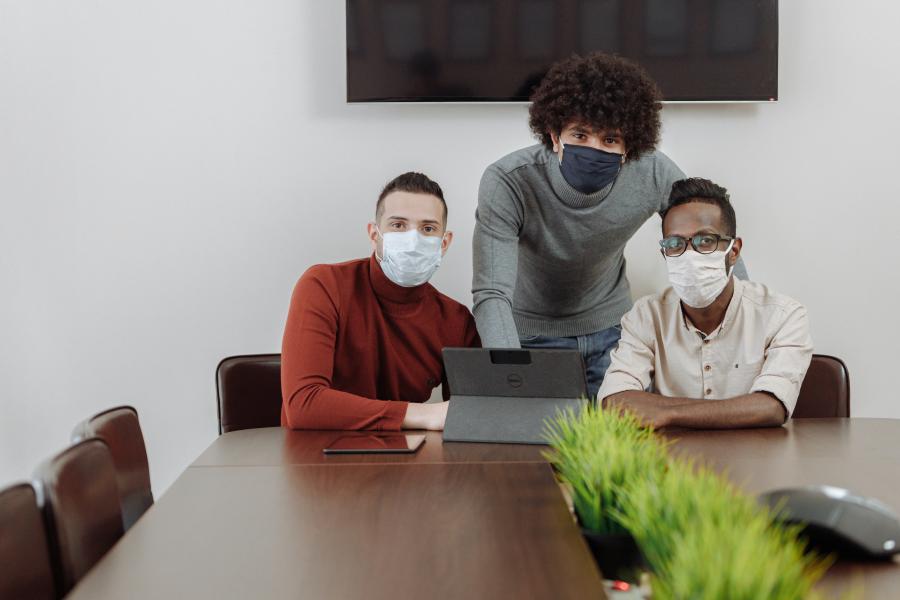 Three men, wearing surgical masks, sitting at a conference table with a laptop.