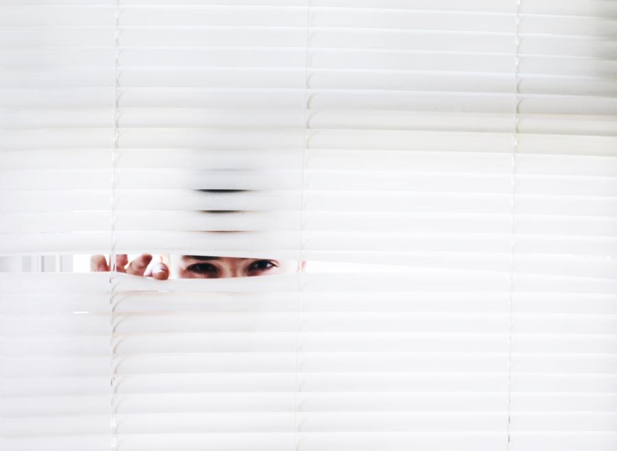 A person's eyes peeking through a wall of white horizontal window blinds