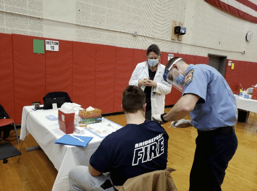 A man in a black BFD t-shirt sits in a school gym while a man in a blue shirt gives him a vaccine