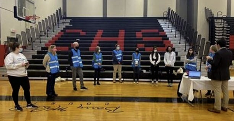 Volunteers in face masks and blue vests stand in a semi-circle in a school gym next to bleachers
