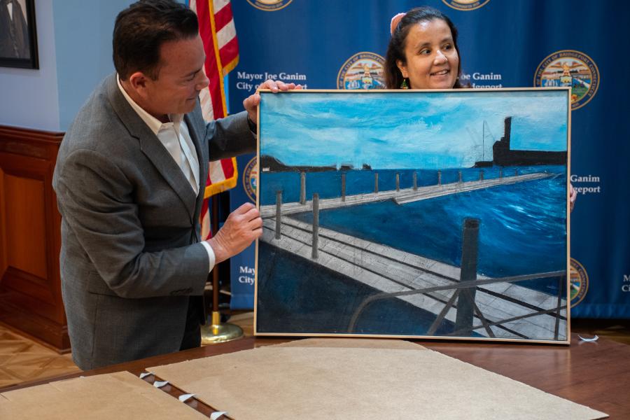 Photo of Mayor Ganim holding a painting while standing next to the painter who presented the piece to him in the Mayor's back conference room. The artist is from The Kennedy Collective in Bridgeport.