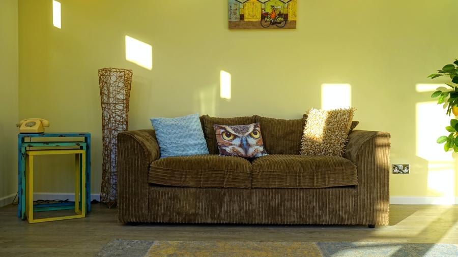 Worn corduroy couch with various pillows in a sunlit yellow room.